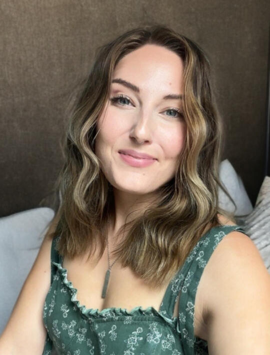 Kate – Still Soft, Still Showing Up A warm portrait of a woman in a green floral dress sitting in soft natural light. She has shoulder-length wavy hair and a gentle smile, with a linen-toned background and cozy, sunset-toned shadows. The photo conveys calm presence and emotional openness.