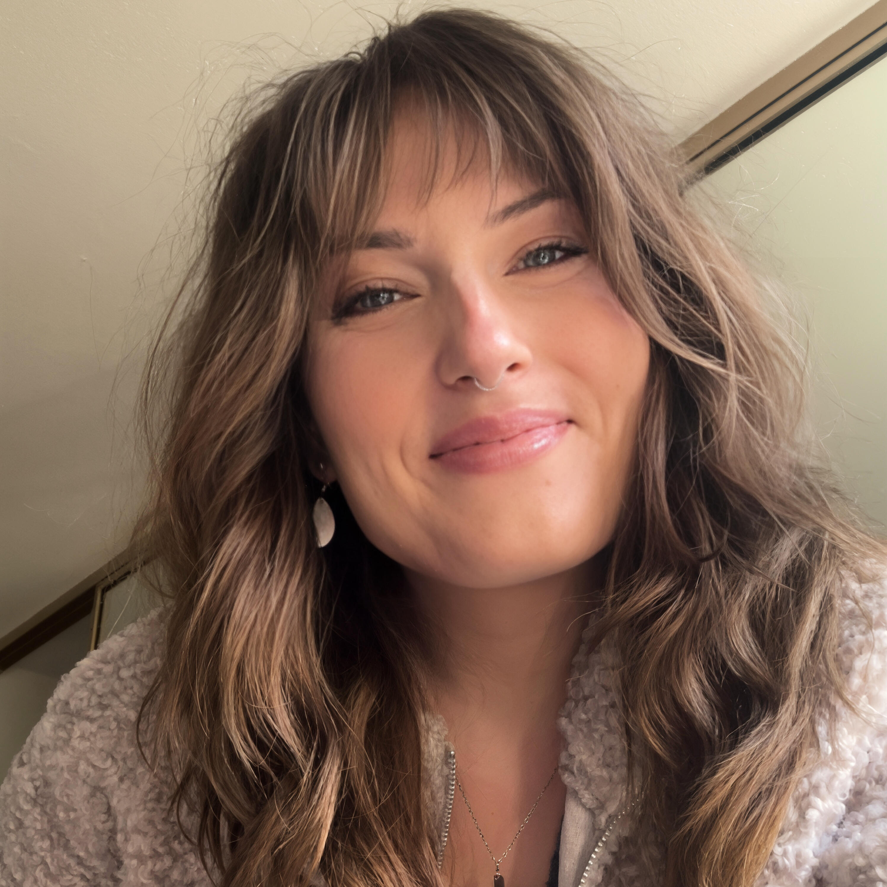 Kate – Still Soft, Still Showing Up A warm portrait of a woman in a green floral dress sitting in soft natural light. She has shoulder-length wavy hair and a gentle smile, with a linen-toned background and cozy, sunset-toned shadows. The photo conveys calm presence and emotional openness.